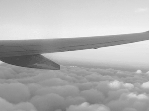 black and white image of wing of flying passengar plane over cloudy sky