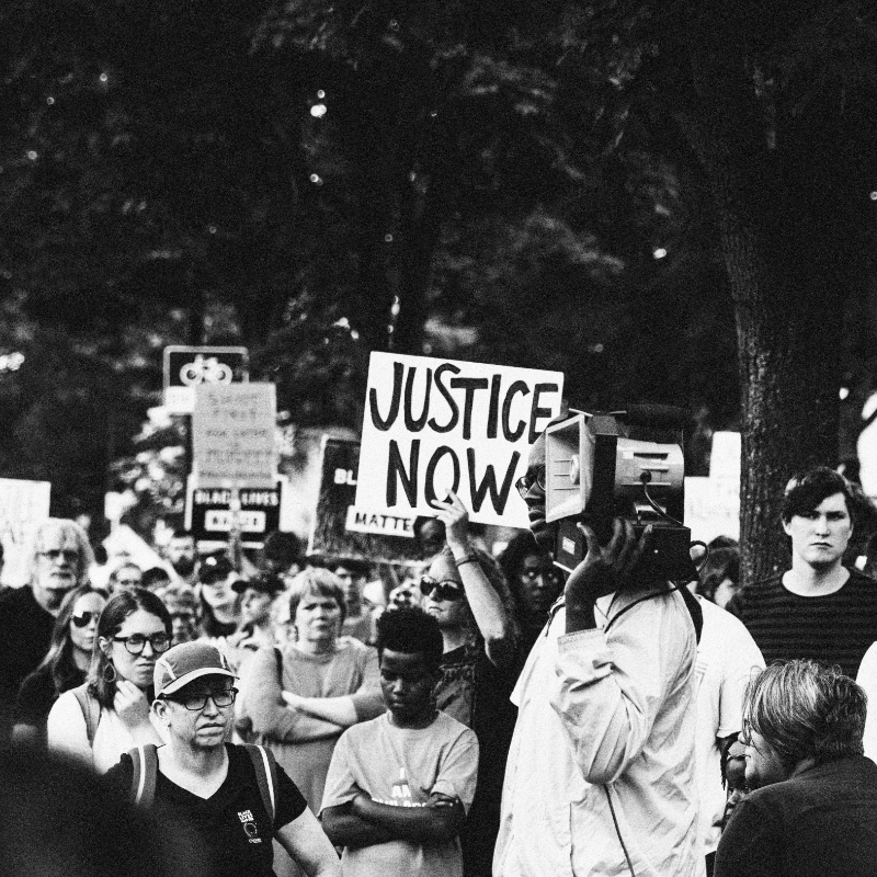A group of people holding placards that read "Justice Now" listening to a black woman speaking at a rally under big trees